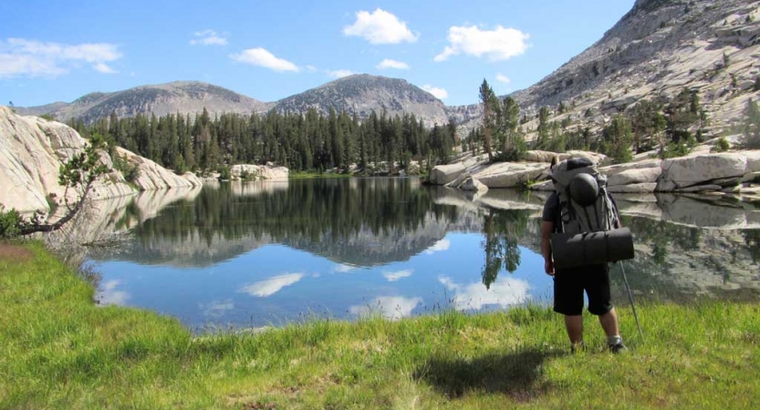 A person wearing a backpack faces away from the camera looking out at an alpine lake. The lake is reflecting the mountains and trees surrounding it. 